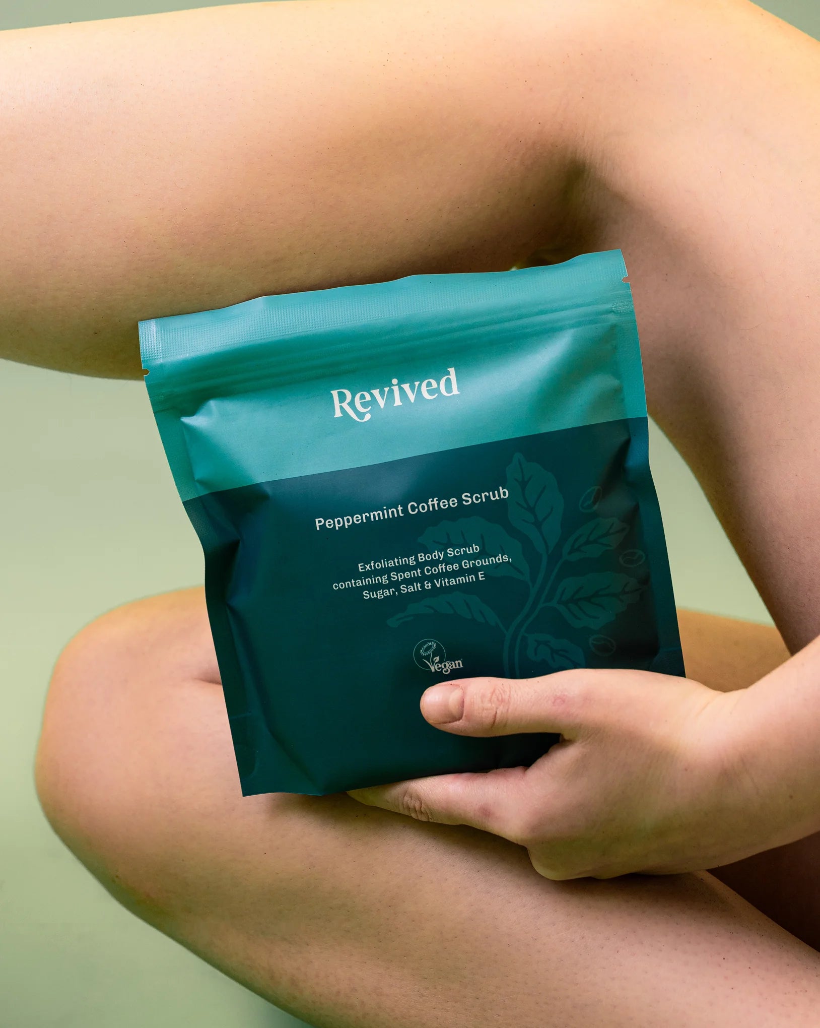Person holding a teal package of Revived Peppermint Coffee Scrub against a light green background