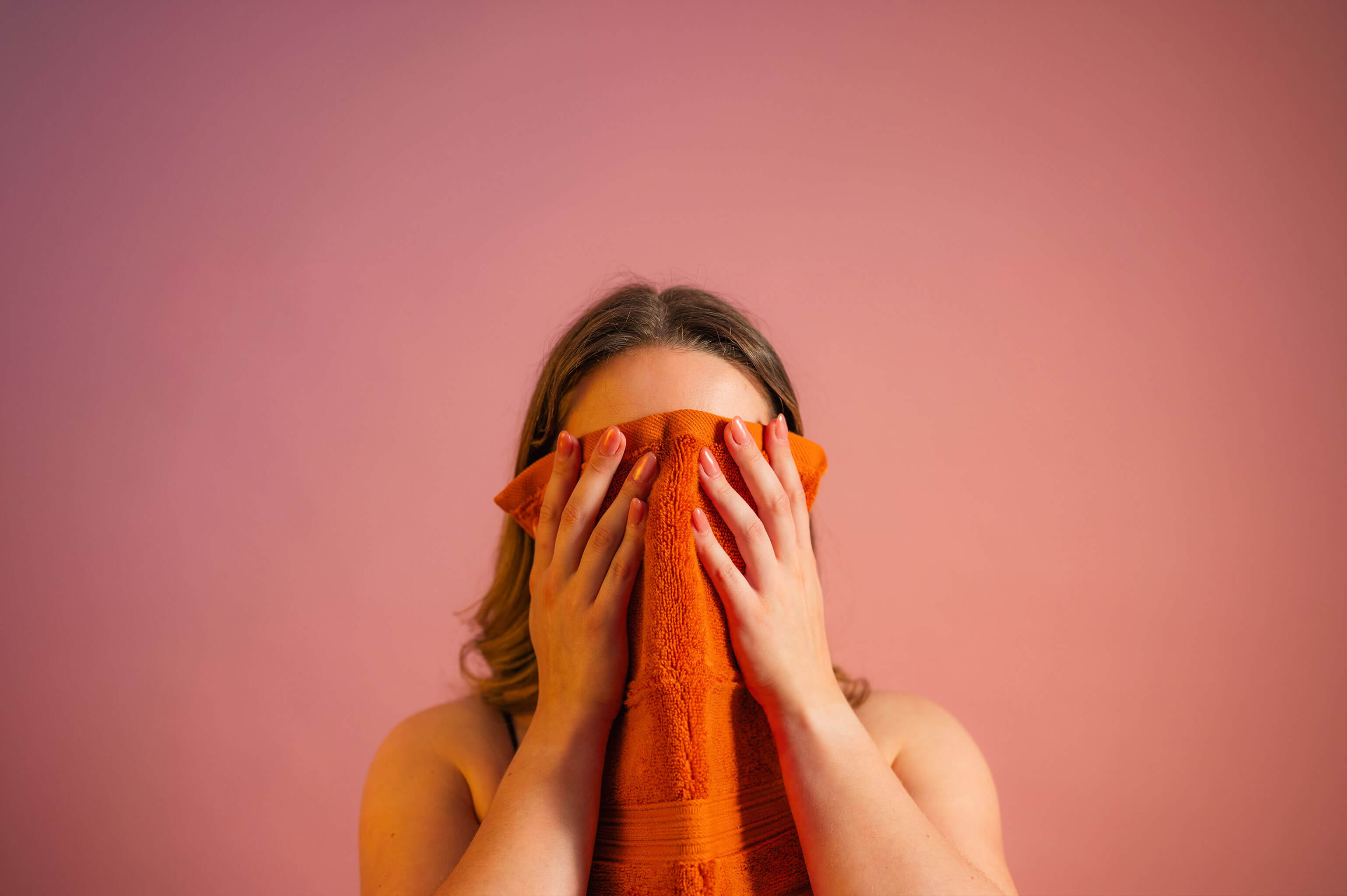 Person covering their face with an orange towel against a pink background