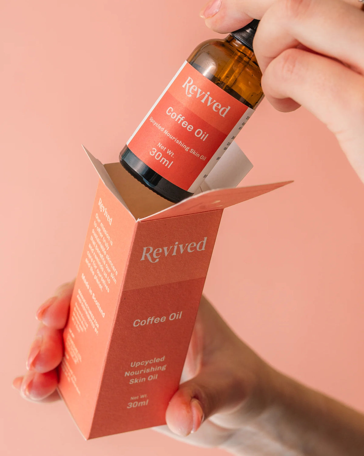 Hand holding a bottle of Revived Coffee Oil with its packaging against a pink background