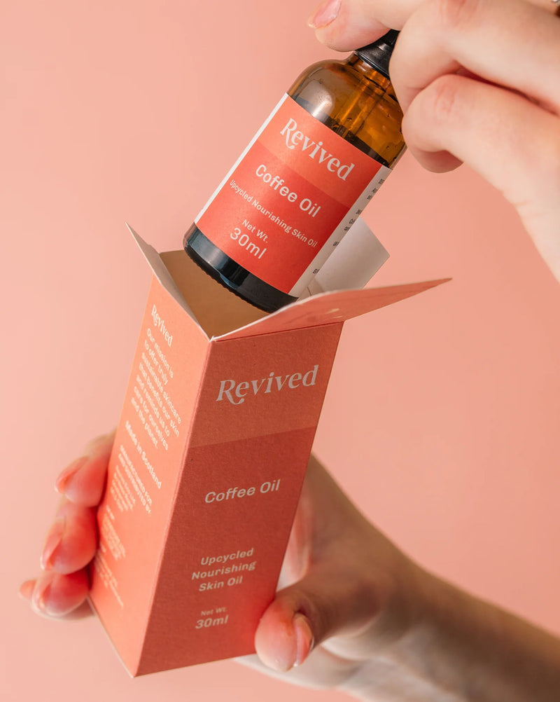 Hand holding a bottle of Revived Coffee Oil with its packaging against a pink background