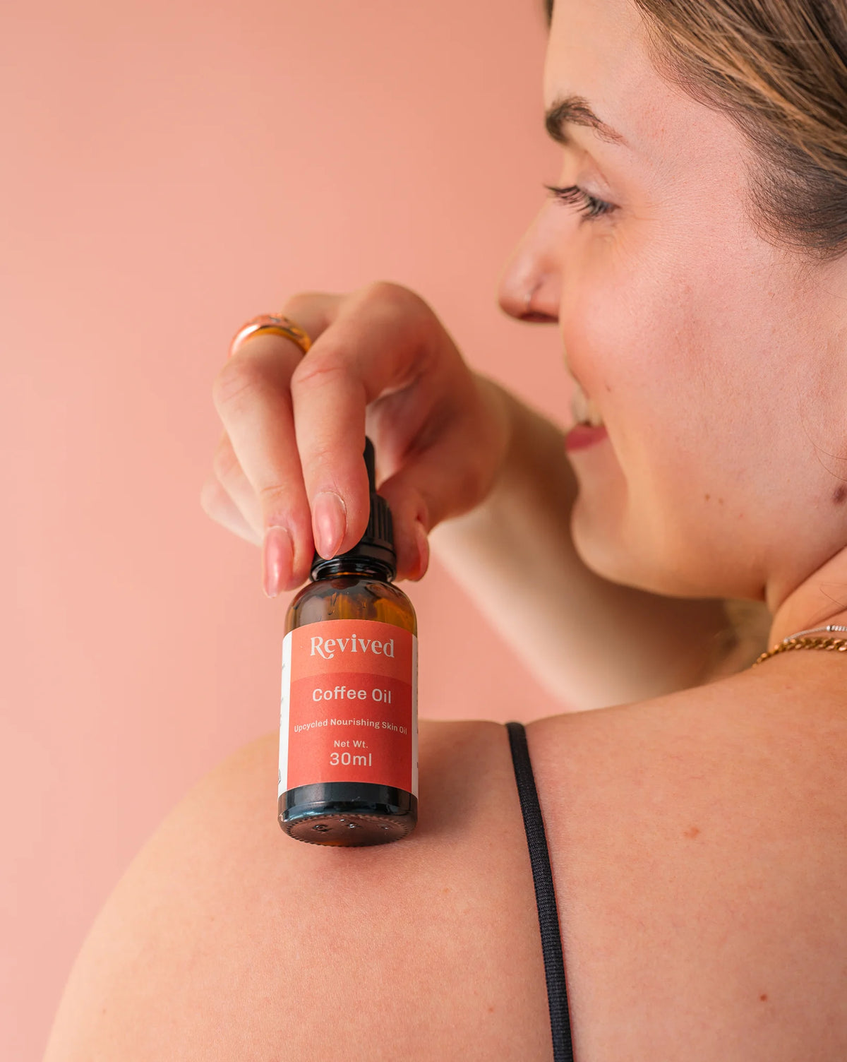 Woman holding a bottle of Revived Coffee Oil over her shoulder against a pink background