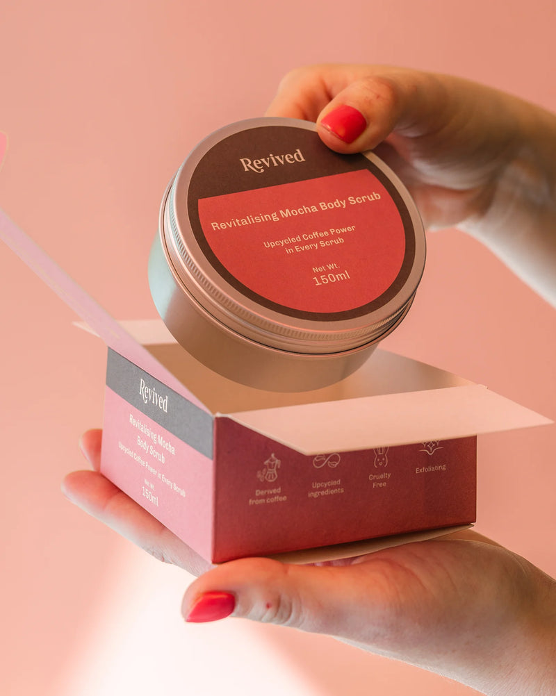 Revived Mocha Body Scrub jar and box held by a hand with red nail polish on a pink background