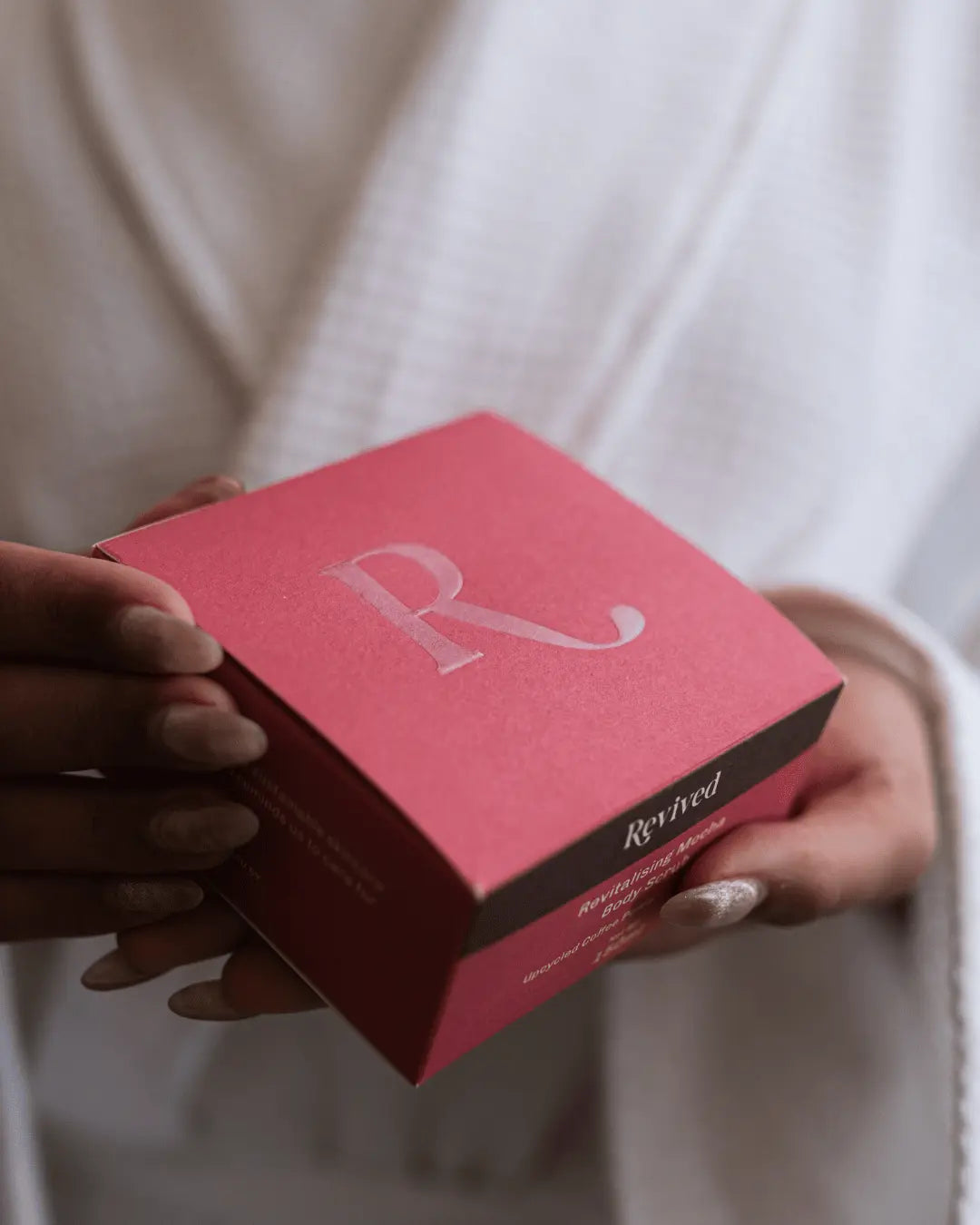 A person in a white robe holds a square pink box of Revived Whipped Mocha Coffee Scrub with a large silver letter R on the lid and the brand name 'Revived' printed on the side.