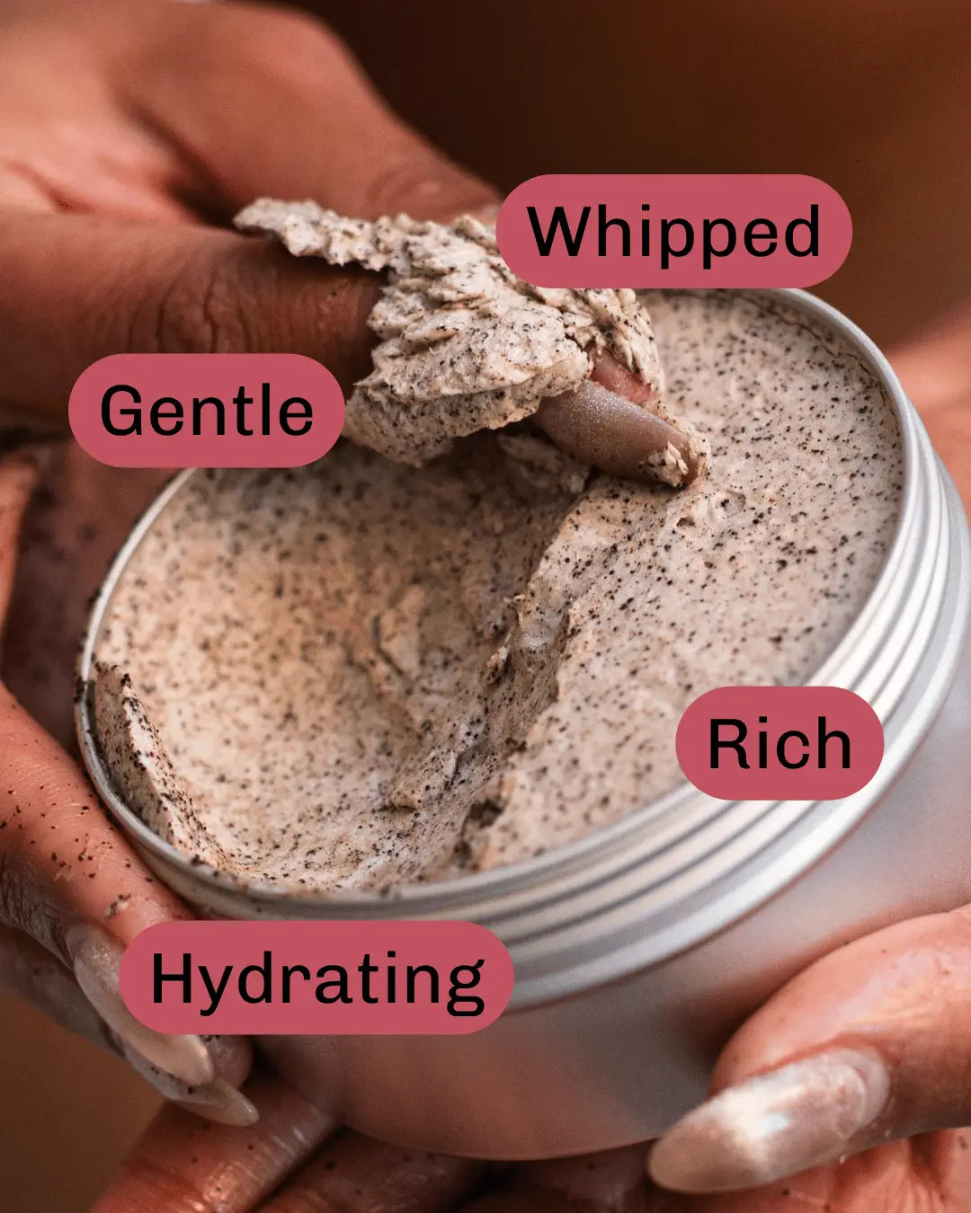 An open aluminium metal jar of whipped mocha exfoliating body scrub being held by hands, with a hand lifting a thick textured layer of the cream product. Pink labels with white text overlay the jar, reading 'Whipped', 'Gentle', 'Rich', and 'Hydrating'.