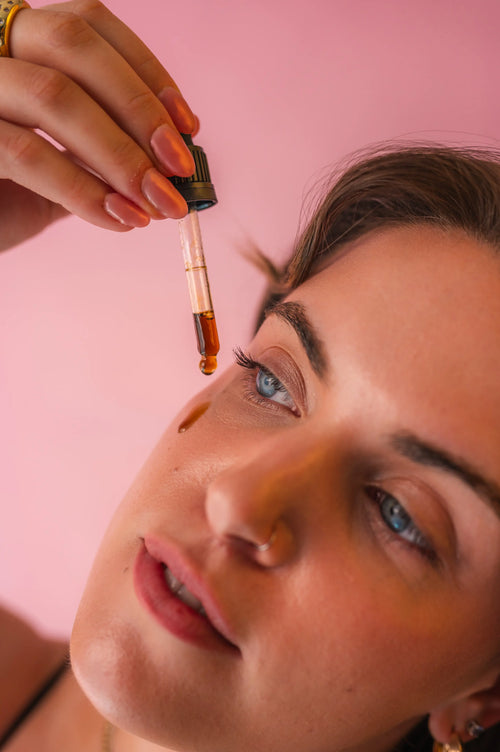 Close-up of a woman applying a dropper with Revived coffee oil to their eye against a pink background