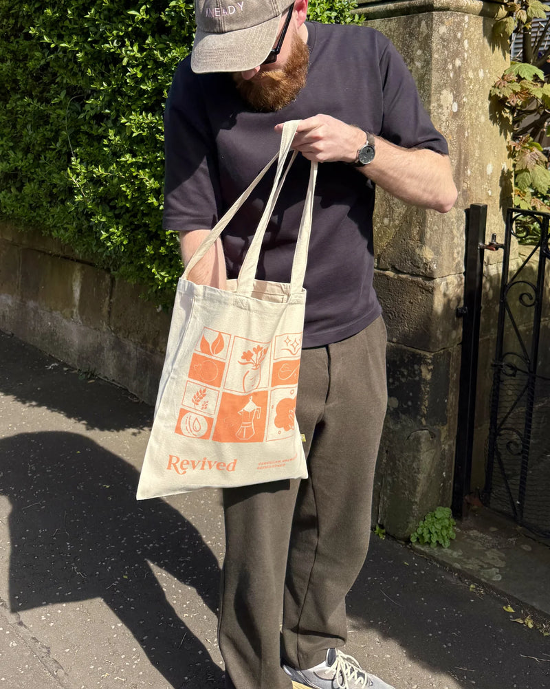 Person holding a tote bag with mosaic graphics and text, standing outdoors near a stone wall.