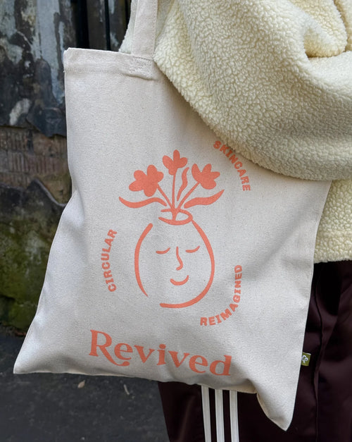 Beige tote bag with 'Revived' wellbeing icon branding held by a person wearing a cream jacket.