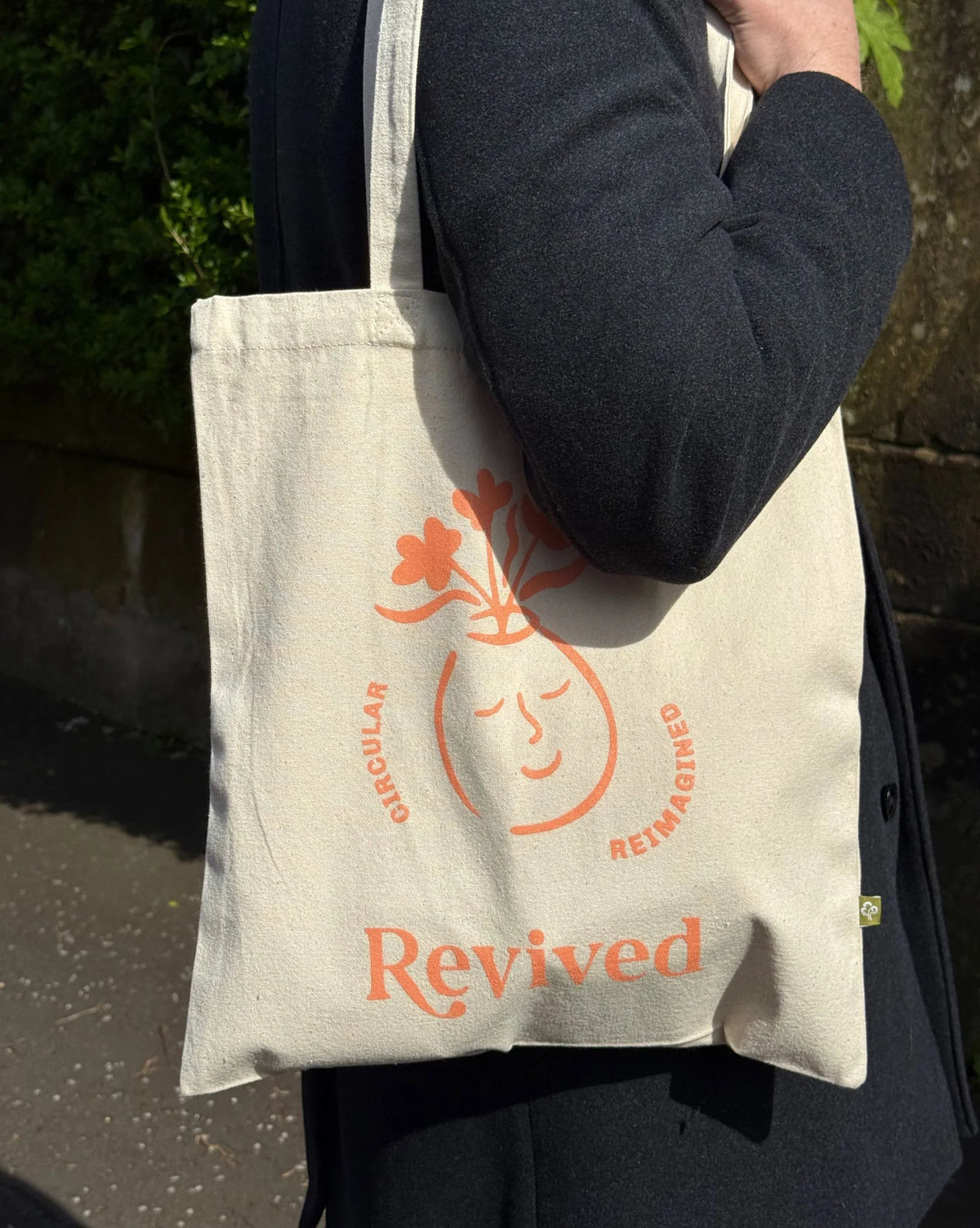 Tote bag with 'Revived' wellbeing icon branding held by a person outdoors.