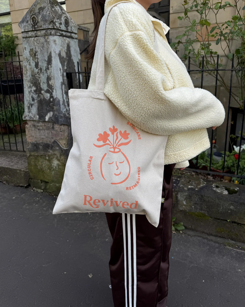 Person holding a  tote bag with 'Revived' wellbeing branding in an outdoor setting.