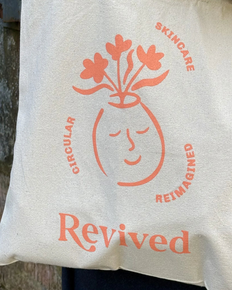 White tote bag with 'Revived' wellbeing branding featuring a face and flowers design.